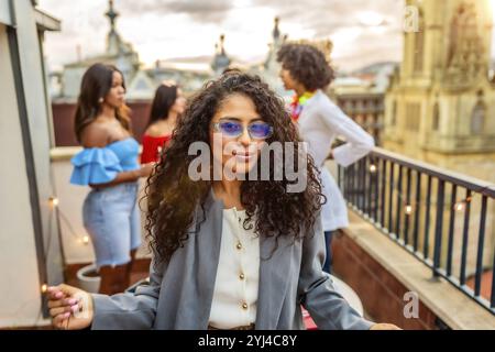 Cool woman in sunglasses smiling while friends partying on the background during rooftop evening party Stock Photo