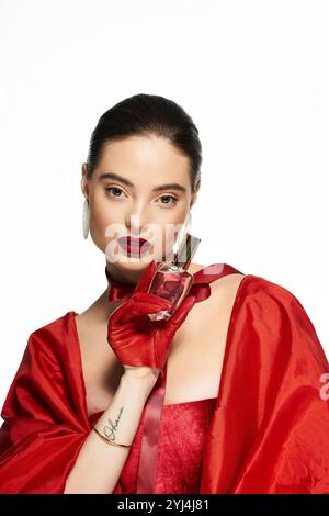 A stunning woman showcases a perfume bottle while dressed in luxurious red fabric. Stock Photo