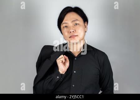 The portrait of a young Asian man wearing the tidy black suit with plain expression on the white grey background shot in the studio. Stock Photo