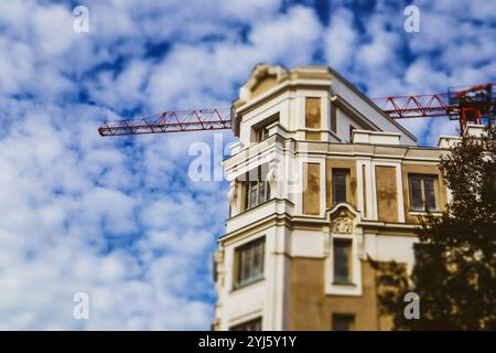 An old beige house, apartment building under construction on a city street. Construction crane machine over a roof and a blue cloudy sky. Restoration Stock Photo