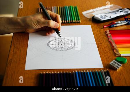 Hand of a Caucasian woman drawing a mandala pattern on a white paper lying on a wooden table, colorful pencils and other drawing utensils nearby Stock Photo