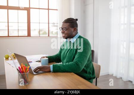 A black guy wearing a green sweater and glasses is working diligently on a laptop at a sunny desk. Nearby, there is a mug and some colorful pens, crea Stock Photo