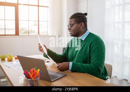 A black man dressed in a green sweater examines printed documents at a table in a well-lit room. His laptop is open, and he pays close attention to th Stock Photo