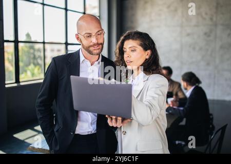 Two colleagues engage in a collaborative discussion while reviewing a laptop. The backdrop features a contemporary office with a large window and a te Stock Photo