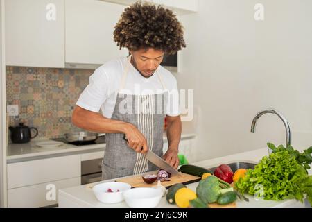 A black man is skillfully chopping vegetables on a wooden cutting board in a well-lit kitchen, surrounded by fresh produce. He is focused on preparing Stock Photo