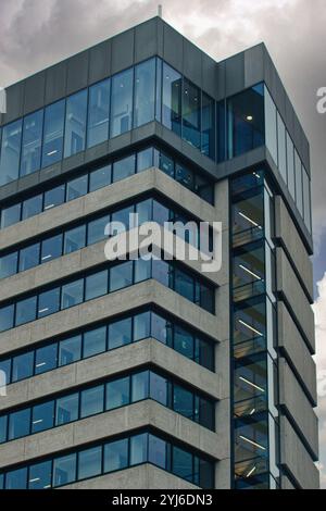 A modern, grey concrete office building with large windows and an internal glass elevator shaft. The building is angled toward the viewer, revealing m Stock Photo