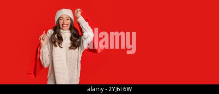 A cheerful young woman dressed in a warm sweater and hat holds shopping bags high in the air, smiling widely against a vibrant red background, embodyi Stock Photo