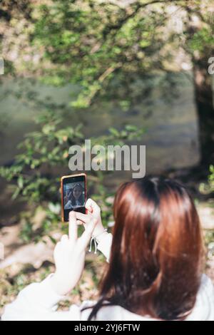 A woman with red hair is seen from behind, taking a selfie with her smartphone while standing near a river in a forested area. Stock Photo