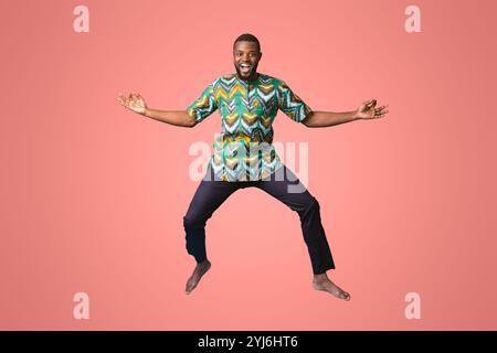 Carefree black man in african costume posing while jumping up on blue studio background. Smiling african american guy wearing traditional clothes, ful Stock Photo