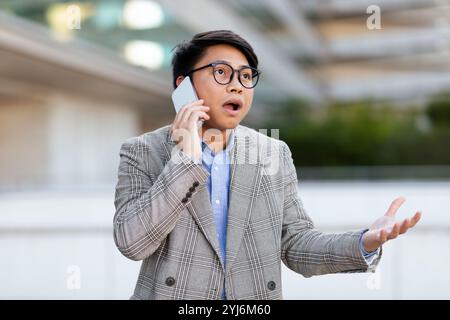 A young Asian man is standing outside, wearing a stylish blazer while talking on his phone and looking surprised. The background features modern archi Stock Photo