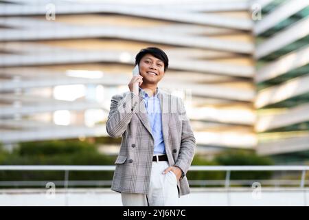 A young Asian man stands confidently in a tailored suit while engaging in a phone conversation outside a contemporary architectural structure during d Stock Photo