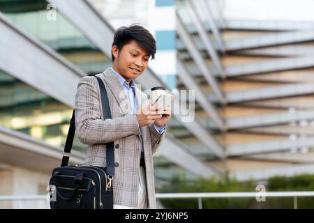 A young Asian businessman in a plaid suit and carrying a bag smiles as he checks his smartphone outside a contemporary office building during the day. Stock Photo