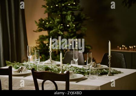 Wooden table with empty polished plates and crystal glasses with cute pine garland in center located in dining room decorated for X-mas party Stock Photo