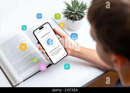 Student studying with smartphone displaying trophy for goal achievement, symbolizing progress tracking and gamification of tasks, with conceptual icon Stock Photo