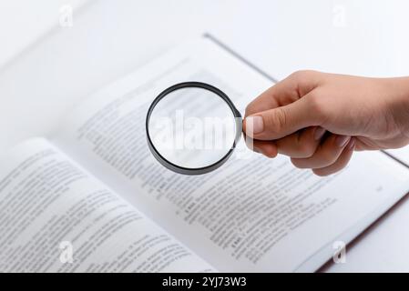 Hand holding magnifying glass searching through open book with dense text, symbolizing research, knowledge seeking, and in-depth reading or study Stock Photo