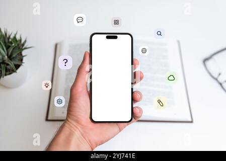 Phone mockup in hand in front of open book with flying icons: question mark, idea, cloud, pen, atom, brain. Great for learning, tech, education visual Stock Photo
