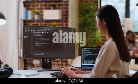 Asian woman is seen typing code for an app development project on her computer, working from a home office. IT specialist engineer focuses on software programming and remote tech work. Camera B. Stock Photo