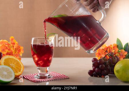 Close-up of glass carafe with homemade summer wine filling a glass surrounded by oranges, lemons and grapes on ceramic table against light brown backg Stock Photo