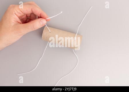 hand is holding toilet paper cardboard roll tube while the other hand is threading a string through holes, part of DIY project Stock Photo