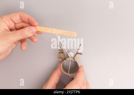 a close-up of a craft project in progress. One hand is holding a popsicle stick with a piece of white string tied around it, other hand is holding a c Stock Photo