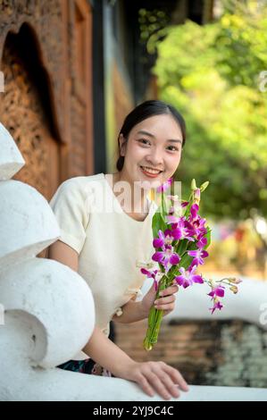A charming Asian woman in a traditional Northern Thai dress stands by the temple, holding a flower and smiling at the camera. Buddhism, spirituality, Stock Photo
