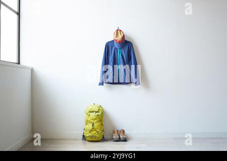 Climbing clothes hanging on white wall by window with backpack and trekking shoes placed under it Stock Photo