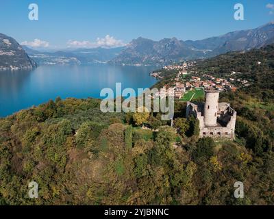 Aerial view of a historic stone tower on a lush hillside overlooking a serene blue lake, surrounded by mountains and a charming lakeside village. Stock Photo