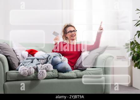 A woman sits comfortably on her couch at home, using her smart glasses to browse the internet for Christmas shopping, while using her credit card to m Stock Photo