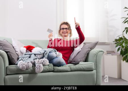A woman sits comfortably on her couch at home, using her smart glasses to browse the internet for Christmas shopping, while using her credit card to m Stock Photo