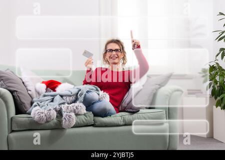 A woman sits comfortably on her couch at home, using her smart glasses to browse the internet for Christmas shopping, while using her credit card to m Stock Photo