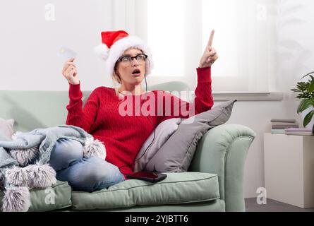 A woman sits comfortably on her couch at home, using her smart glasses to browse the internet for Christmas shopping, while using her credit card to m Stock Photo