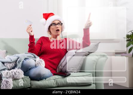 A woman sits comfortably on her couch at home, using her smart glasses to browse the internet for Christmas shopping, while using her credit card to m Stock Photo
