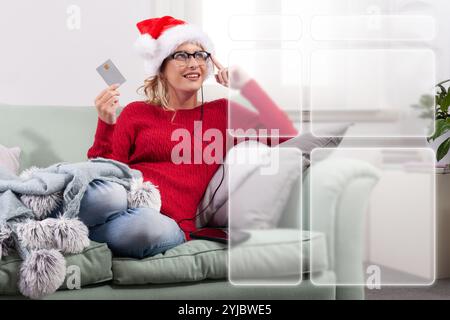 A woman sits comfortably on her couch at home, using her smart glasses to browse the internet for Christmas shopping, while using her credit card to m Stock Photo