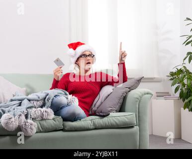 A woman sits comfortably on her couch at home, using her smart glasses to browse the internet for Christmas shopping, while using her credit card to m Stock Photo