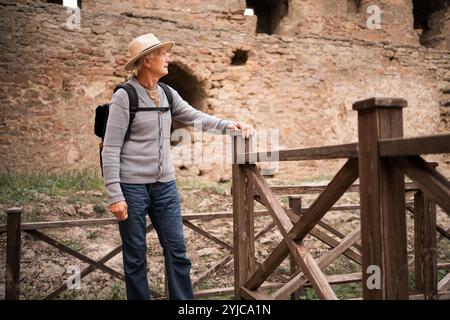 Elderly man with backpack and hat exploring ancient fortress, standing near wooden fence, looking around, wearing casual outfit, old stone walls in Stock Photo