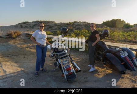 Two women stand beside their motorcycles, soaking in the warm sunset glow against the sandy backdrop. Stock Photo