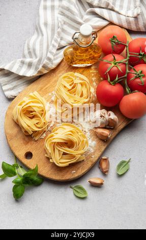 Uncooked tagliatelle pasta nests on wooden cutting board, tomatoes, garlic, basil and olive oil, italian food concept Stock Photo