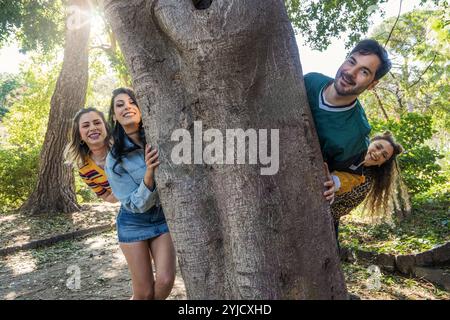 Cheerful multiethnic group of friends having fun while hiding behind large tree during sunny afternoon in urban park - People lifestyle concept Stock Photo