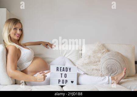 Young woman, blonde, pregnant, 9 months, lying on sofa, pointing at sign READY TO POP, smiling, Stuttgart, Baden-Wuerttemberg, Germany, Europe Stock Photo