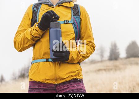 Waist up woman hiker looking away from the camera holding a blue thermo flask in her hands, hiking on a foggy autumn day Stock Photo