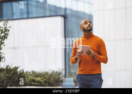 Caucasian man in an orange sweater standing outside in an urban setting holding a digital device, office buildings in the background Stock Photo