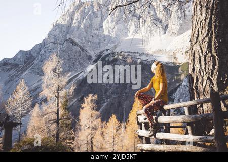 Side view of young caucasian woman hiker in colorful clothes, hiking gear, sitting on a wooden fence somewhere up in the mountains, enjoying the view Stock Photo