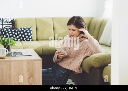 Young caucasian woman with long brown hair in pastel pink sweater and jeans working from home on her laptop and phone. Cheerful woman student studying Stock Photo