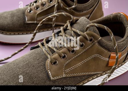 Laces and eyelets of khaki canvas sneakers close-up. Pair of new brown textile shoes with white soles and lacing over pale purple background. Stock Photo