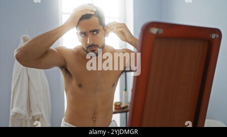 Young hispanic man with a beard examines his reflection in a spa room with calm and focused expression, highlighting grooming and wellness in a serene Stock Photo