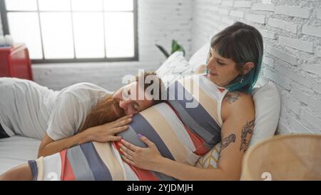 Pregnant woman relaxing on bed while her partner listens to her belly, symbolizing love and family bond in the cozy setting of their home bedroom. Stock Photo