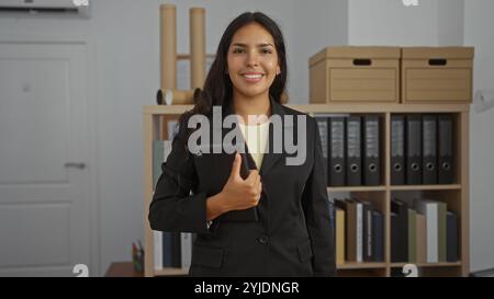 Young woman in office workplace smiles while holding a tablet in a professional environment with shelves and binders in the background. Stock Photo