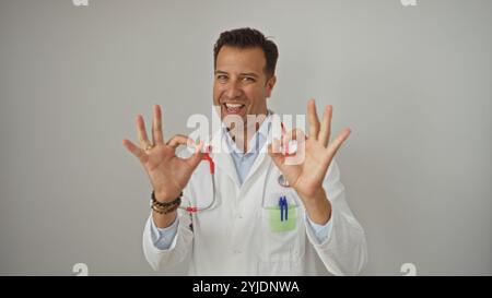 Handsome hispanic man in a white coat makes an ok gesture with both hands while smiling over an isolated white background Stock Photo