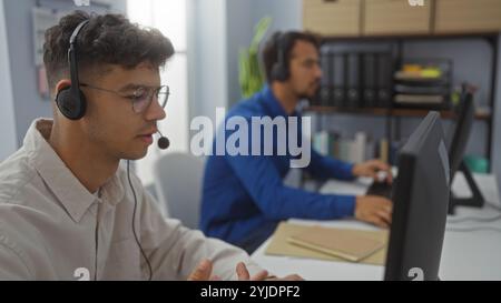 Hispanic men working together in an office, wearing headsets in a business environment, focused on their tasks and collaborating as employees or execu Stock Photo