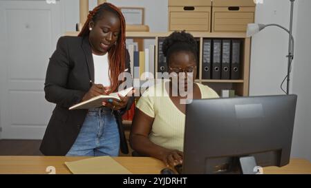 African american women business partners working together indoors in an office, with one writing in a notebook and the other typing at a computer. Stock Photo